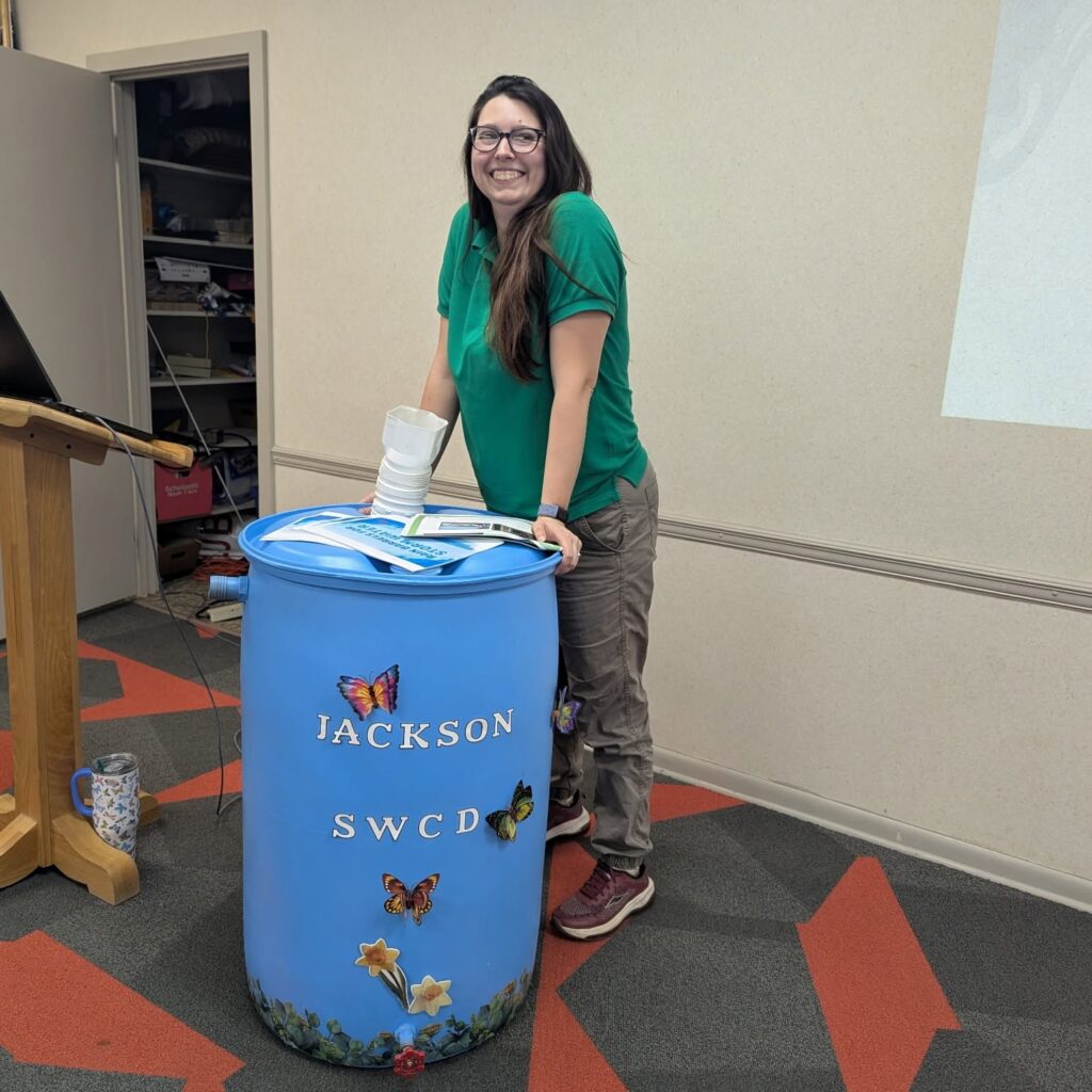 Ag Specialist, Ashley Hansen, in a green JSWCD polo, smiling and leaning on a blue, decorated rain barrel at the rain barrel workshop